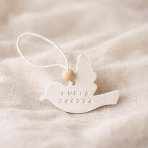 Puede incluir: Adorno en forma de paloma de cerámica blanca con el nombre "LUCIA" y la fecha "14.08.24" en relieve. Cuenta con una cuenta de madera y una cuerda para colgar. El adorno se encuentra sobre un fondo neutro y texturizado.