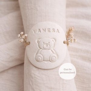 Könnte beinhalten: Ein weißer, ovaler Serviettenring mit dem eingeprägten Namen "VANESA" oben und einem Teddybären-Design darunter. Der Ring ist mit kleinen Blumen und Bindfaden verziert. Der Text "Can be personalised" ist ebenfalls sichtbar.