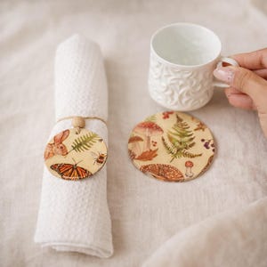 Puede incluir: Un rollo de servilleta blanco con una etiqueta de madera con un diseño de conejo, mariposa, abeja y helecho. También hay un posavasos a juego y una taza de té blanca con un estampado floral. El posavasos tiene un diámetro de unos 10 cm.