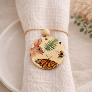 Può includere: Un portatovagliolo rotondo in legno con un disegno dipinto raffigurante un coniglio, una farfalla monarca, un'ape e una felce. L'anello è legato con spago e ha una perla di legno. Il portatovagliolo è avvolto attorno a un tovagliolo di lino bianco.