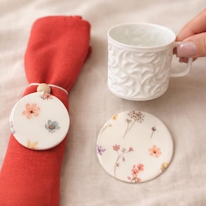 Puede incluir: Una taza de té de cerámica blanca con un diseño floral texturizado es sostenida por una mano. Una servilleta roja se sujeta con un servilletero con decoración floral. Una posavasos floral a juego está cerca. Los diseños florales incluyen flores rosas, naranjas y azules.
