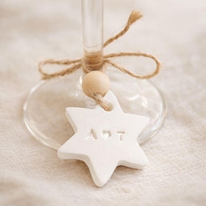 Puede incluir: Una copa de vino transparente adornada con una etiqueta blanca en forma de estrella. La etiqueta, con la inscripción "A ♡ T", está suspendida por un lazo de cuerda y una cuenta de madera. La copa descansa sobre una tela de color neutro.