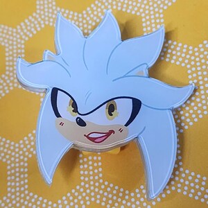 May include: A silver and white cartoon character with a mischievous grin. The character has yellow eyes and a black nose. The character is a pin or button.