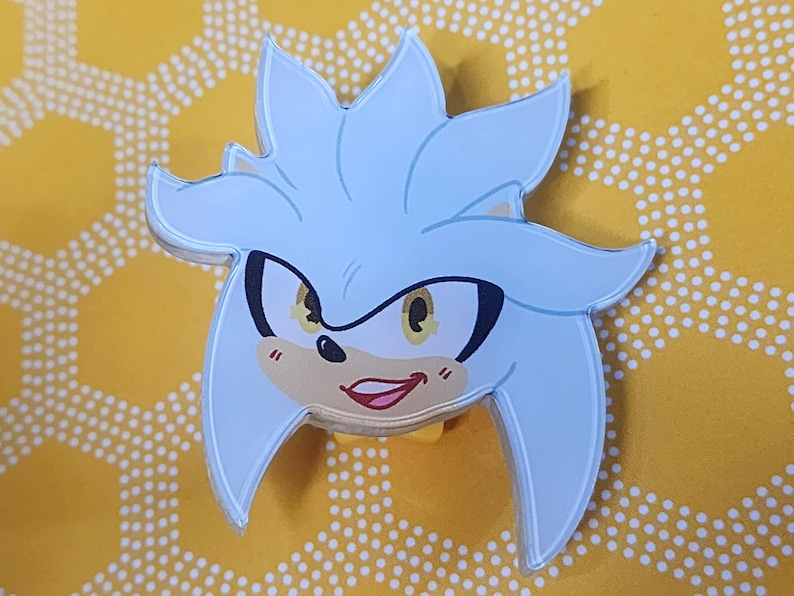 May include: A silver and white cartoon character with a mischievous grin. The character has yellow eyes and a black nose. The character is a pin or button.