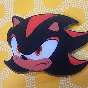 May include: A black and red cartoon character with yellow eyes and a red nose. The character has a serious expression and is looking to the right. The character is a pin or button.