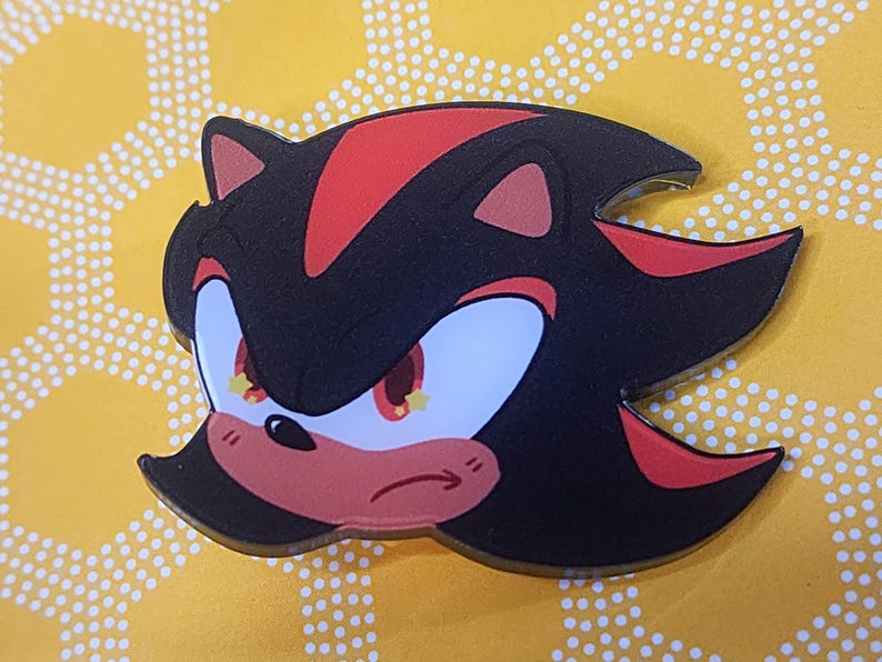 May include: A black and red cartoon character with yellow eyes and a red nose. The character has a serious expression and is looking to the right. The character is a pin or button.