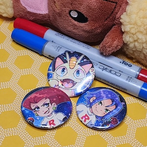 May include: Three round button pins with a space-themed design. The pins feature a cartoon illustration of a cat-like Pokemon, a female character with red hair, and a male character with blue hair. The pins are on a yellow background with a honeycomb pattern.
