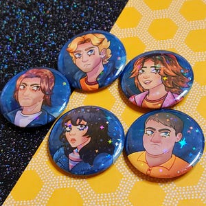 Peut inclure: Cinq boutons ronds bleus et blancs avec une finition scintillante. Chaque bouton présente un portrait de personnage de dessin animé avec une coiffure et une expression différentes. L'arrière-plan de chaque bouton est bleu foncé avec des étoiles blanches.