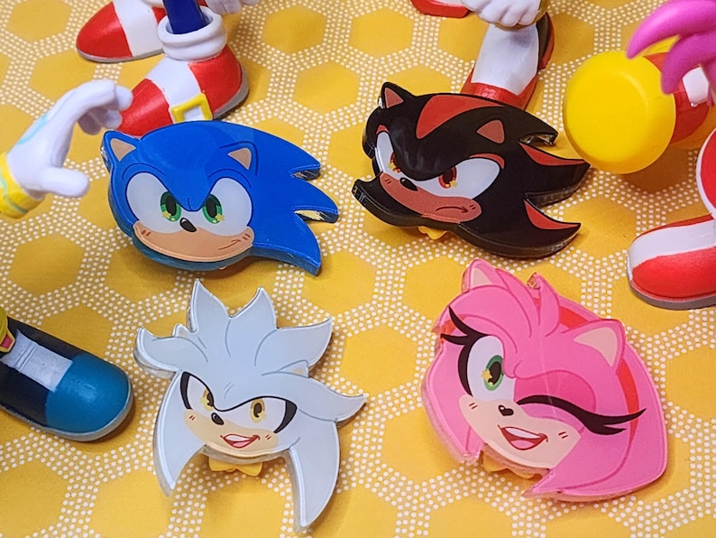 May include: Four colorful cartoon character pins featuring Sonic the Hedgehog, Shadow the Hedgehog, Amy Rose, and Silver the Hedgehog. The pins are made of plastic and have a glossy finish.