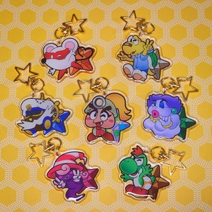 Könnte beinhalten: Acht bunte Charakter-Schlüsselanhänger mit goldenen Stern-Anhängern. Die Charaktere umfassen eine lila und blaue Wolke, einen grünen Dinosaurier, einen pink- und blauen Charakter mit einem Werkzeug, einen lila Charakter mit einem Hut, eine weiße Maus mit einer roten Maske, einen braunen Charakter mit einem blauen Hut und einem Steuerrad, einen gelben Charakter mit einem blauen Hut und einem Werkzeug und einen gelben Charakter mit einem blauen Hemd und einer grünen Muschel.