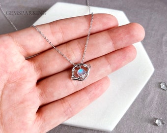 Puede incluir: Un collar de plata con un colgante de piedra lunar en forma de planeta rodeado por un anillo de plata con pequeñas estrellas. El collar está en una mano.