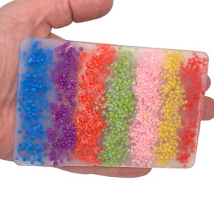 Op de afbeelding: Een rechthoekig, transparant blok van hars met ingebedde gekleurde kralen. De kralen zijn gerangschikt in horizontale strepen in de kleuren blauw, paars, oranje, groen, roze en geel.