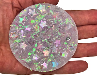 May include: A clear round coaster with iridescent stars and green glitter embedded in the resin.