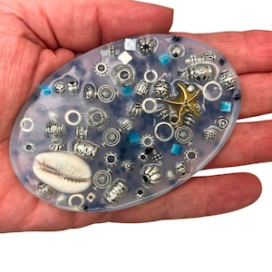 Op de afbeelding: Een heldere ovale harsbak gevuld met zilveren en gouden kralen, een schelp en een gouden zeester. De bak ligt op een hand.