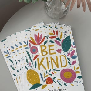 May include: A stack of coloring books with a white background and colorful floral designs. The cover features the text "Be Kind" in gold lettering and the words "Positive Affirmation Coloring Book".