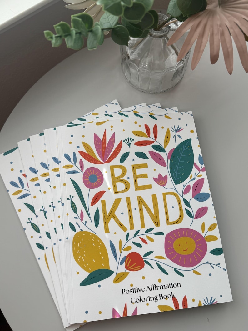 May include: A stack of coloring books with a white background and colorful floral designs. The cover features the text "Be Kind" in gold lettering and the words "Positive Affirmation Coloring Book".