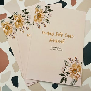 May include: A stack of pink self-care journals with floral designs and the text "30-day Self Care Journal" and "includes guide and daily prompts".