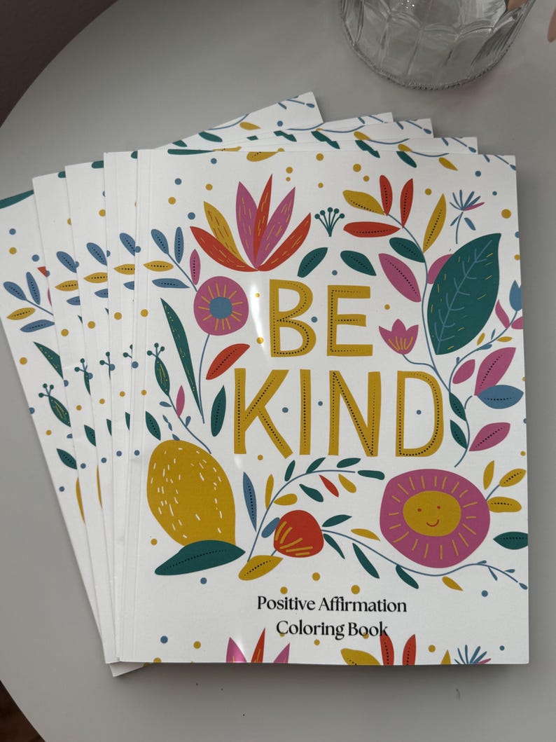 May include: A stack of coloring books with a white background and colorful floral designs. The cover features the text "Be Kind" in gold lettering and the words "Positive Affirmation Coloring Book".