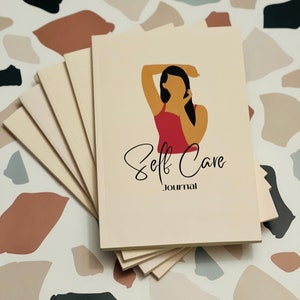 May include: A stack of cream-colored journals with the words "Self Care Journal" on the cover. The cover features an illustration of a person with their arm raised. The background is a terrazzo pattern.