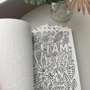 May include: An open coloring book with a page featuring the words "I AM Strong" surrounded by floral and leaf designs. The opposite page is blank.