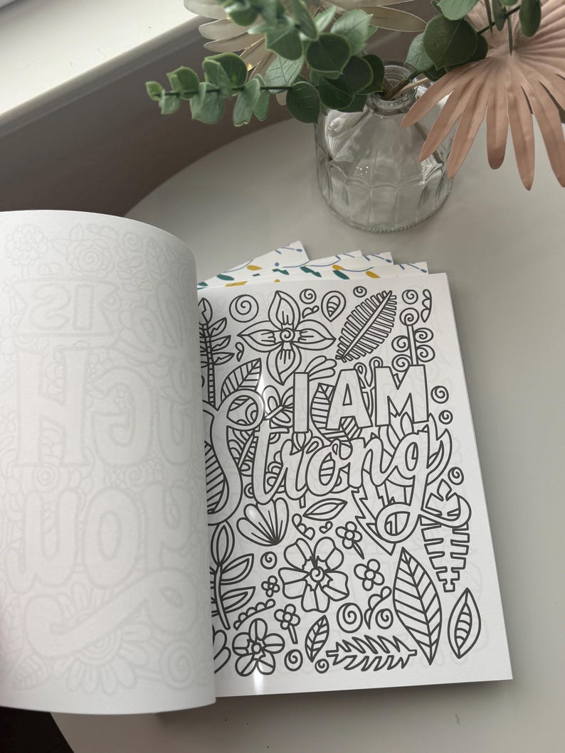 May include: An open coloring book with a page featuring the words "I AM Strong" surrounded by floral and leaf designs. The opposite page is blank.