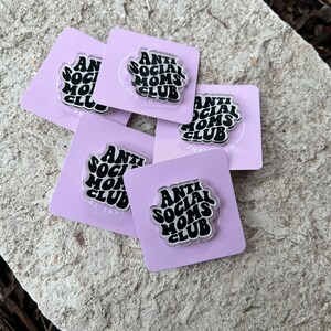 May include: Five purple cards with the text "Anti Social Moms Club" in black on a clear acrylic pin. The pins are on a gray stone surface.
