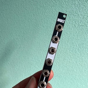 Puede incluir: Módulo Eurorack blanco y negro con el texto "MULT" y un logotipo de oso. El módulo tiene siete conectores plateados.
