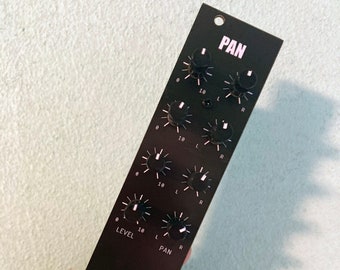 May include: A black electronic audio module with the word "PAN" printed at the top. It features multiple knobs, input jacks, and the text "LEVEL". The module has a bear logo and is held in a hand.