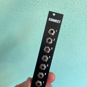 Puede incluir: Panel de metal negro con la palabra "CONNECT" impresa en blanco. El panel tiene ocho conectores de color plateado con los números del 1 al 8 impresos junto a ellos. Un logotipo de oso blanco se encuentra en la parte inferior del panel.