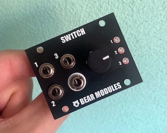 May include: A black electronic music module with the text "SW1TCH" and "BEAR MODULES" printed on it. The module has three knobs and a rotary knob with a white line.