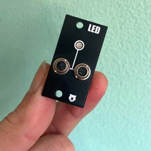 Puede incluir: Módulo electrónico rectangular negro con dos conectores redondos plateados y un indicador LED blanco. El módulo tiene un logotipo de oso blanco y el texto "LED" impreso en él.