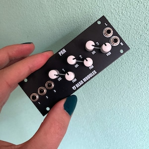 Puede incluir: Un módulo de sintetizador negro Bear Modules con tres botones etiquetados "LVL" y "PAN" y tres conectores de 6,35 mm etiquetados "1", "2" y "3". El módulo tiene dos conectores de 6,35 mm etiquetados "L" y "R" en la esquina superior derecha. El módulo está etiquetado "BEAR MODULES" en texto blanco.