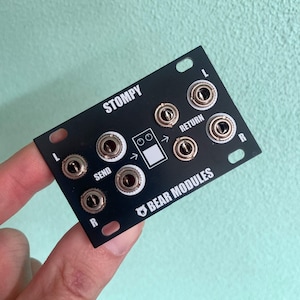 Puede incluir: Pedal de efectos de audio negro Stompy Bear Modules con múltiples conectores de entrada y salida. El pedal tiene el texto "STOMPY" y "BEAR MODULES" impreso. El pedal tiene un gráfico blanco en el centro con flechas.
