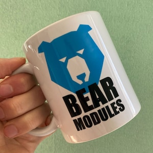 Puede incluir: Taza de cerámica blanca con un gráfico de oso azul y las palabras "BEAR MODULES" en negro. La taza tiene un asa curva. El gráfico del oso es un diseño geométrico. Fondo verde claro.