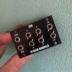 Puede incluir: Módulo Eurorack negro con el texto "DIV" y "BEAR MODULES" impreso en él. El módulo tiene seis conectores plateados con LED rojos a su lado. Los conectores están etiquetados como "CLOCK", "1/2", "1/4", "1/8", "1/16" y "1/32".