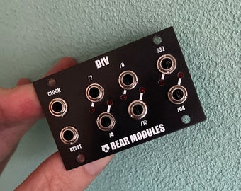 May include: Black Eurorack module with the text "DIV" and "BEAR MODULES" printed on it. The module has six silver jacks with red LEDs next to them. The jacks are labeled "CLOCK", "1/2", "1/4", "1/8", "1/16", and "1/32".