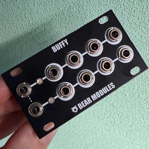 May include: A black electronic module with the word "BUFFY" at the top and "BEAR MODULES" at the bottom. It features multiple silver and black connectors arranged in rows. The module has mounting holes at each corner.