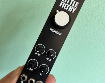 May include: Black electronic module with white text that reads "LITTLE FILTHY" and "FREQUENCY". It has a large white knob and several smaller black knobs labeled "LP-HP", "RESO", "~VE", and "CV". The module also has input and output jacks.