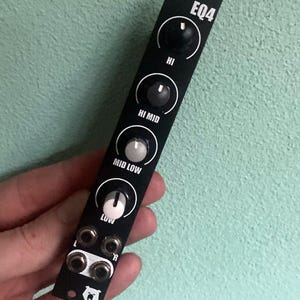 Puede incluir: Módulo de ecualizador de 4 bandas negro con perillas etiquetadas como "HI", "HI MID", "MID LOW" y "LOW". El módulo tiene dos conectores de 6,35 mm etiquetados como "L" y "R" y un logotipo de oso blanco.