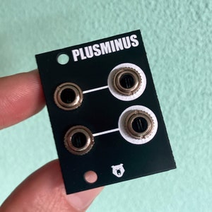 Puede incluir: Módulo negro con el texto "PLUSMINUS" y cuatro jacks de color dorado. Un logotipo de oso blanco se encuentra en la parte inferior central.