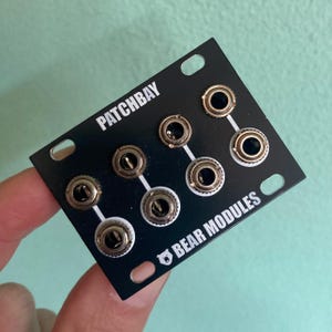 Puede incluir: Módulo electrónico negro con el texto "PATCHBAY" y "BEAR MODULES". El módulo tiene varios conectores circulares con detalles plateados y dorados. El módulo es rectangular con esquinas redondeadas y orificios de montaje.