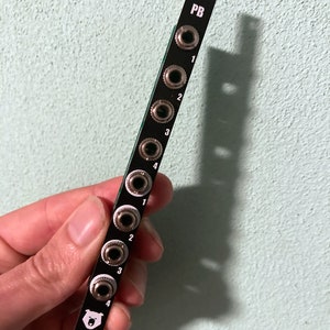 Puede incluir: Patch bay negro con ocho conectores jack de 1/4 de pulgada y el texto "PB" impreso en la parte superior. El patch bay tiene un logotipo de oso blanco impreso en la parte inferior.