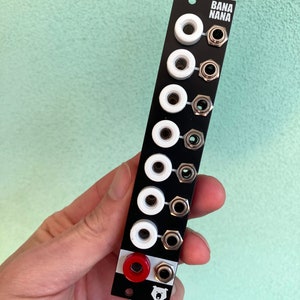 Puede incluir: Panel negro con ocho jacks blancos y un jack rojo. El panel tiene el texto "BANA NANA" impreso en él. Un pequeño logotipo de oso está en la parte inferior del panel.