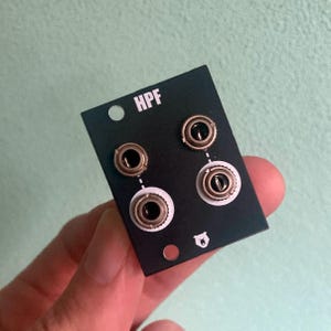 Könnte beinhalten: Schwarzes Panel mit dem Text "HPF" und vier goldfarbenen Metallbuchsen. Das Panel hat ein kleines Bären-Symbol in der unteren rechten Ecke.