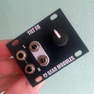 Puede incluir: Módulo electrónico negro con la inscripción "TILT EQ" y "BEAR MODULES". Cuenta con una perilla con un indicador blanco, cuatro conectores de entrada y un conector blanco. El módulo tiene orificios de montaje en cada esquina.