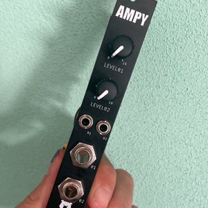Puede incluir: Interfaz de audio de metal negro con el texto "AMPY" impreso en blanco. Tiene dos perillas etiquetadas "LEVEL 01" y "LEVEL 02" con marcas blancas. Hay cuatro conectores de audio de 1/4 de pulgada etiquetados "01" y "02".