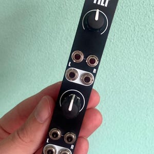 Puede incluir: Módulo electrónico negro con la palabra "TILT" en blanco. Tiene dos perillas y varios conectores de entrada/salida etiquetados como "L" y "R". Un pequeño logotipo de oso en la parte inferior. El módulo se sostiene contra un fondo verde claro.
