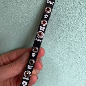 Puede incluir: Panel de metal negro con la palabra "SPLIT" impresa en blanco. El panel tiene seis conectores con las etiquetas: "LEFT TIP", "RIGHT RING", "STEREO", "LEFT TIP", "RIGHT RING", y "STEREO".