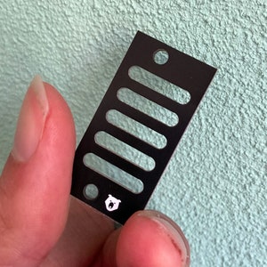 Puede incluir: Placa de metal negro con seis ranuras y un logotipo de oso blanco. La placa está sostenida por una mano.