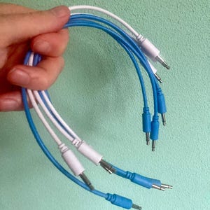May include: A selection of audio cables with 3.5mm jacks. The cables are blue and white, with white and blue connectors. The cables are held in a hand against a light green backdrop. The cables are for audio equipment.